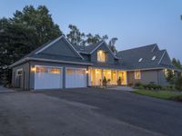 large suburban house with large driveway and garage with lit windows at dusk, next to trees