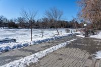 the walkway by the pond is covered in snow and is surrounded by bare trees in winter