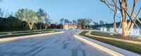 this paved path has trees and fountains along with benches around it on the side of the walkway are tall trees and the buildings are also