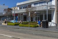 people are walking and riding on the sidewalk near shops in an open area with bushes