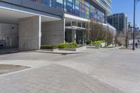 a sidewalk that has bushes on each side of it with buildings in the background and a building across the street