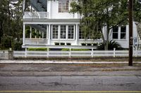 a white fence surrounding a street with plants on it and parked cars behind it in the distance