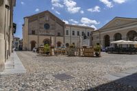 there are many tables on this stone plaza near a building and other buildings with an arch, outside a fountain and on the side,