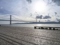 the road by the water is cobble stone and has benches near it with a suspension bridge behind it