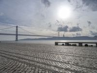 the road by the water is cobble stone and has benches near it with a suspension bridge behind it