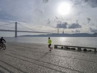 the road by the water is cobble stone and has benches near it with a suspension bridge behind it