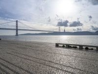 the road by the water is cobble stone and has benches near it with a suspension bridge behind it