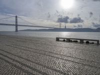 the road by the water is cobble stone and has benches near it with a suspension bridge behind it