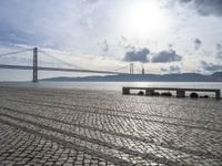 the road by the water is cobble stone and has benches near it with a suspension bridge behind it