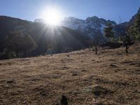 the sun shining over the mountains behind the field and trees in the foreground of the scene