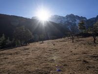 the sun shining over the mountains behind the field and trees in the foreground of the scene