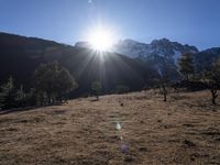 the sun shining over the mountains behind the field and trees in the foreground of the scene