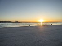 a man riding a skateboard across a sandy beach near the ocean at sunset time