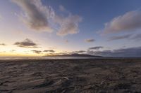 a beach with a lone bench, and clouds on a blue sky, with the sun setting in the distance