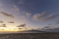 a beach with a lone bench, and clouds on a blue sky, with the sun setting in the distance