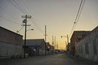 the sun sets behind power lines in an empty alley in a city setting along with a few buildings