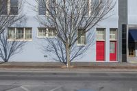 a red door is standing on the sidewalk and behind the fire hydrant is a grey wall with multiple windows and trees