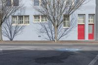 a red door is standing on the sidewalk and behind the fire hydrant is a grey wall with multiple windows and trees