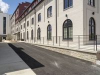 a street with a paved, white sidewalk and long buildings that are behind a fence