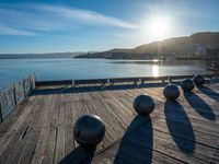 five steel balls are sitting on the pier overlooking water with sun shining over the ocean and city in the background