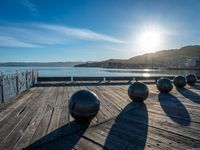 five steel balls are sitting on the pier overlooking water with sun shining over the ocean and city in the background