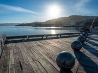 five steel balls are sitting on the pier overlooking water with sun shining over the ocean and city in the background