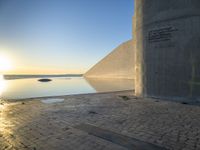 a sunset with sun rising over the water and a large concrete structure on it's side