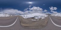 a road curves in a very cloudy sky near mountains with snow on it and a blue sky