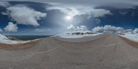 a road curves in a very cloudy sky near mountains with snow on it and a blue sky