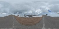 a road curves in a very cloudy sky near mountains with snow on it and a blue sky