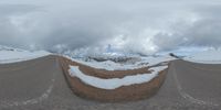 a road curves in a very cloudy sky near mountains with snow on it and a blue sky