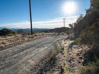 the sun shines behind power lines on a country road near brush and rocks in an arid area