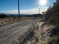 the sun shines behind power lines on a country road near brush and rocks in an arid area