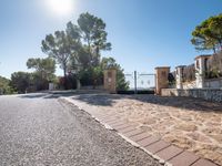 a paved driveway has stone bricks between trees and bushes and a fence and fenced off street area