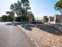 a paved driveway has stone bricks between trees and bushes and a fence and fenced off street area