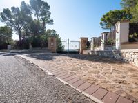 a paved driveway has stone bricks between trees and bushes and a fence and fenced off street area