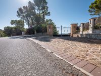 a paved driveway has stone bricks between trees and bushes and a fence and fenced off street area