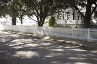 the white fence is in the street beside a white house with trees and bushes on both sides
