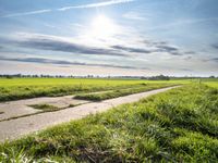 an empty road that is near the grass fields with the sun shining over the field