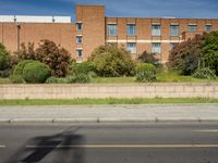 an empty street and a tall brick building with several windows behind a wall along side the road