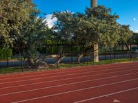 a red running track next to trees and fencing with a clock tower in the distance