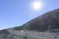 a rainbow colored kite is soaring above the rocky hill side and rocks below it on a sunny day