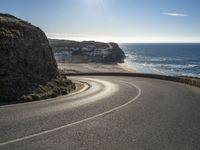 a curved road near the beach with waves coming up in front of the cliff and the sun