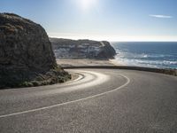 a curved road near the beach with waves coming up in front of the cliff and the sun