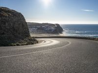 a curved road near the beach with waves coming up in front of the cliff and the sun