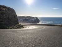 a curved road near the beach with waves coming up in front of the cliff and the sun