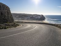 a curved road near the beach with waves coming up in front of the cliff and the sun