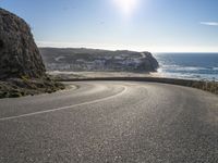 a curved road near the beach with waves coming up in front of the cliff and the sun