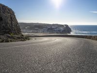 a curved road near the beach with waves coming up in front of the cliff and the sun