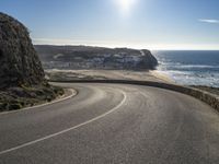 a curved road near the beach with waves coming up in front of the cliff and the sun